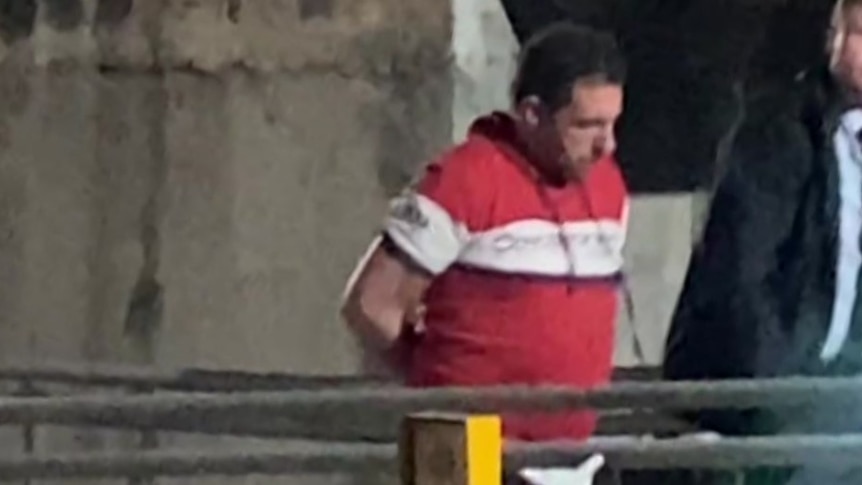 A man in a red and white shirt strides along a concrete walkway in handcuffs with a police officer beside him