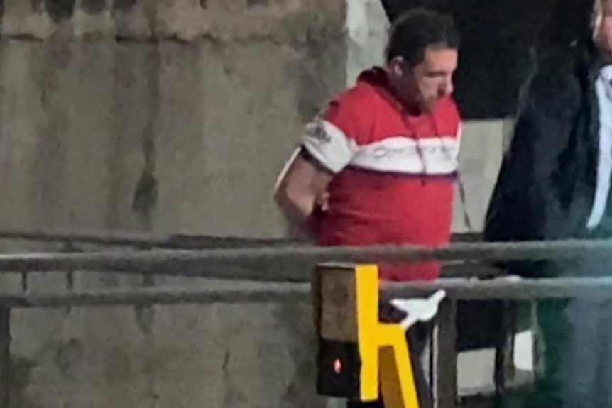 A man in a red and white shirt strides along a concrete walkway in handcuffs with a police officer beside him