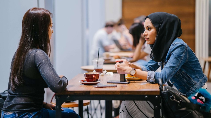 Two women sit at a table in a cafe, talking together.