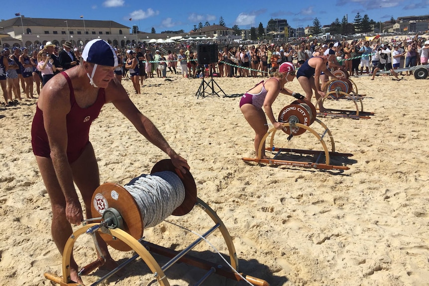 Men and women in 1938 lifesaving costumes man hand-operated winches on Bondi Beach.