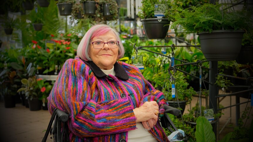 An older woman with multi-coloured hair sitting down in a plant shop 