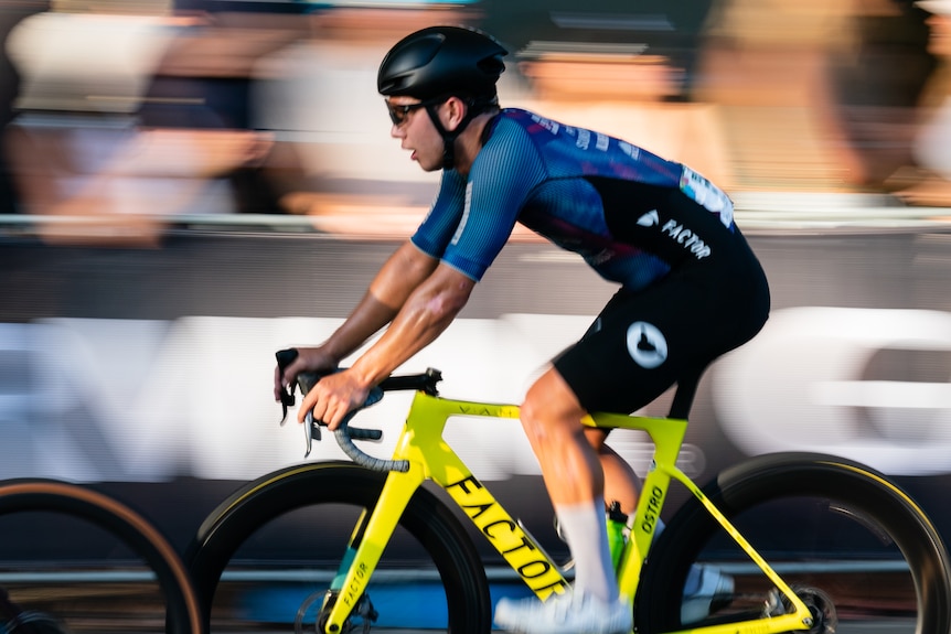 A man wearing blue cycling jersey, helmet, glasses, speeds along on a bike.