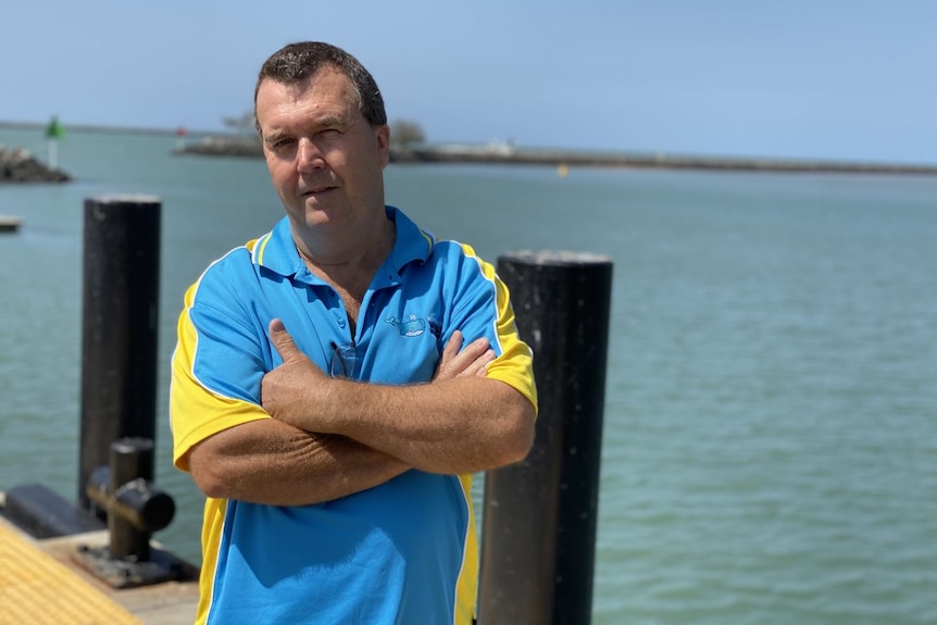 A man, wearing a blue and yellow polo shirt, stand on a jetty with his arms folded