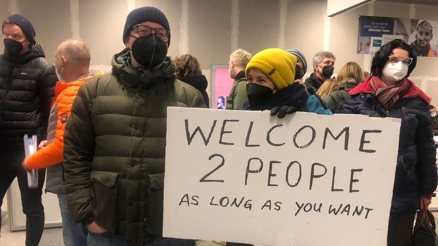A father and son hold up a hand-written cardboard sign saying "Welcome 2 people as long as you want". 
