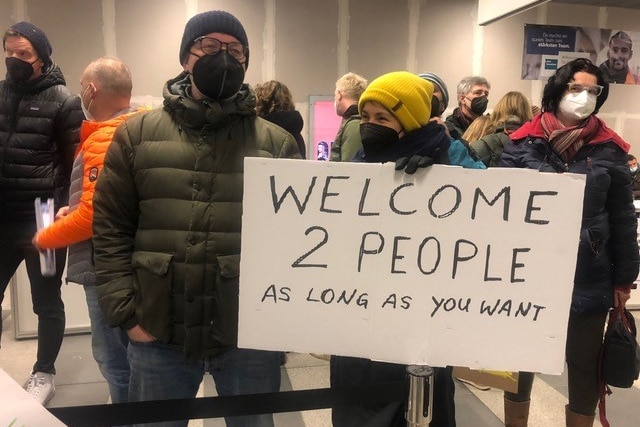 A father and son hold up a hand-written cardboard sign saying "Welcome 2 people as long as you want". 