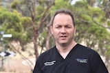 A middle-aged man with receding brown hair wears black scrubs and stands near trees