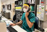 A man wearing green scrubs, a face mask and face shield sits at a desk talking on a landline phone