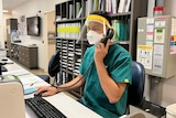 A man wearing green scrubs, a face mask and face shield sits at a desk talking on a landline phone