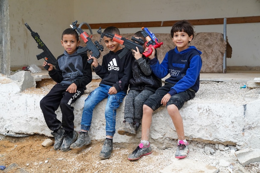 Four young boys holding toy guns and sitting in a ruined building, smiling at the camera