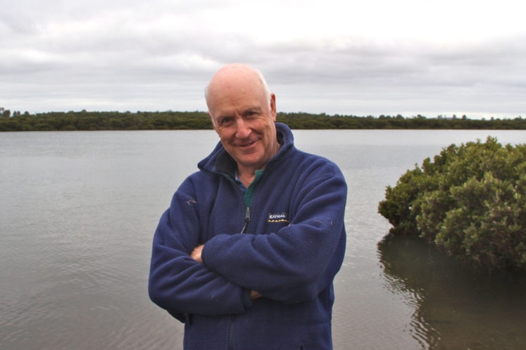 John Clarke stands with his armed crossed in front of an inlet.