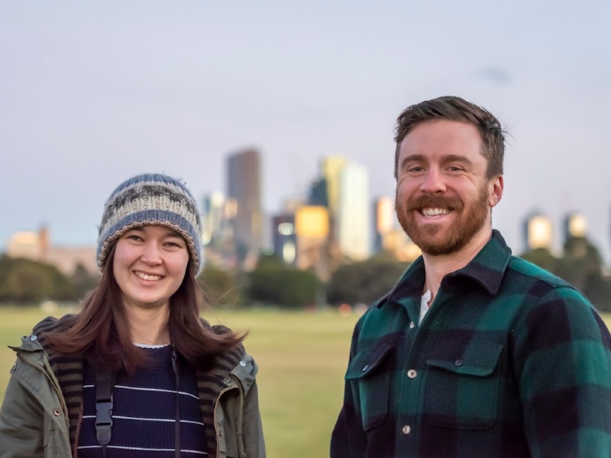 A lady  and a man stand in a park with tall buildings in the background