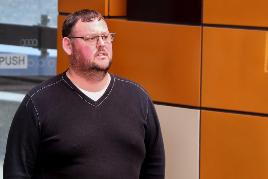 Tim Batchelor, with brown hair, facial hair and glasses, wearing a black jumper, walks past orange Federal Court wall