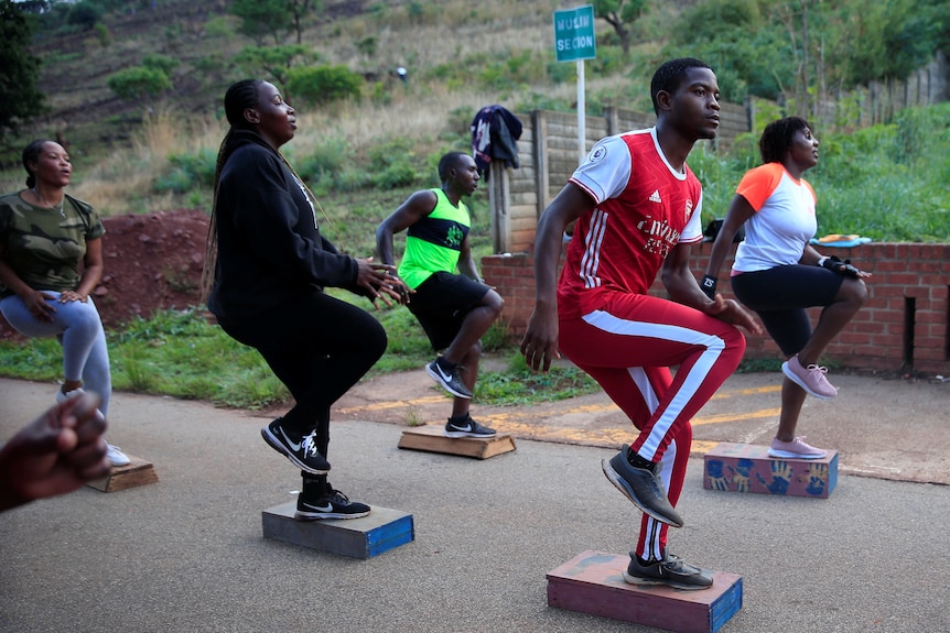 people in exercise clothing lift their knees as they do step ups on raised platforms on a road inside a cemetery