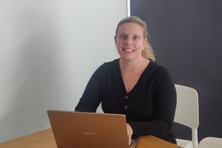 A blond woman with a pontail smiling and sitting at a table with a laptop. 