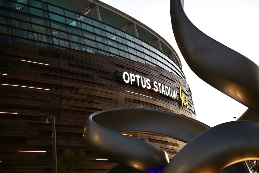 A large squiggly steel art-work partially obscures Perth Stadium at dusk.