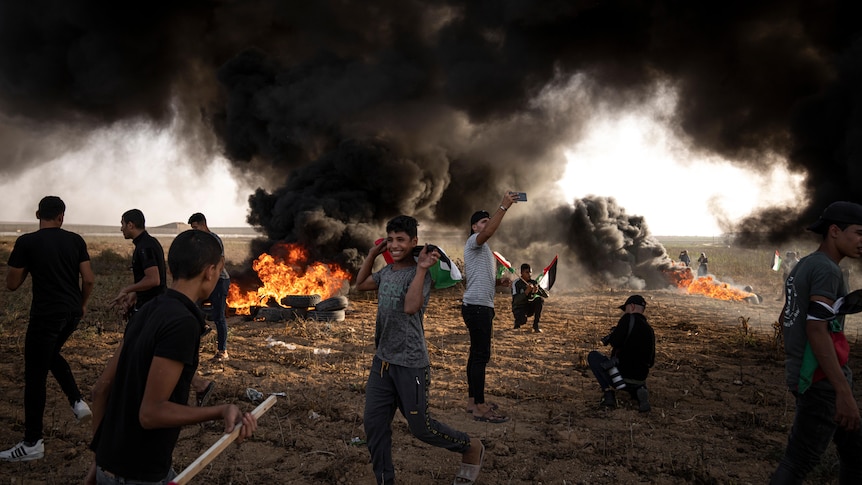 A group of teenagers burn tires on a barren hill, sending thick black smoke into the sky.