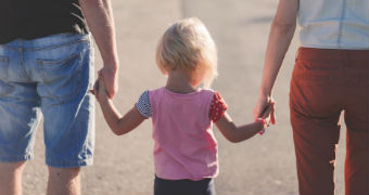 A child holds hands with two adults standing on her left and right.