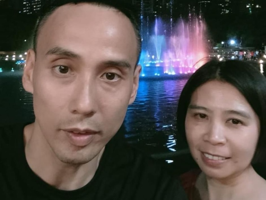 A selfie picture of man and woman at night with colourful lights under a water fountain behind them