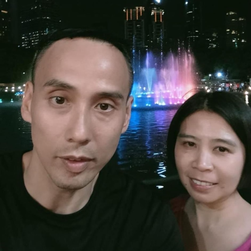 A selfie picture of man and woman at night with colourful lights under a water fountain behind them