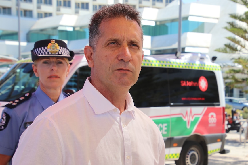 WA Police Minister Paul Papalia listening to a media conference outdoors in front of a policewoman and an ambulance.