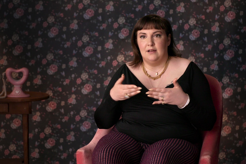 The writer Lena Dunham - a woman in her mid 30s with short fringe and dark top - sits on a chair, gesticulating