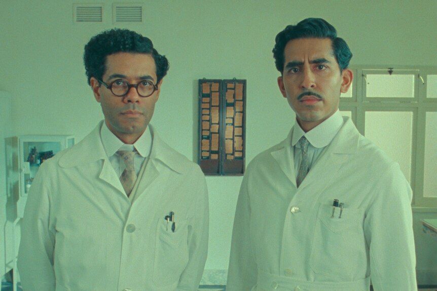 Dev Patel and Richard Ayoade stand in white doctor's coats with slicked back hair looking very serious