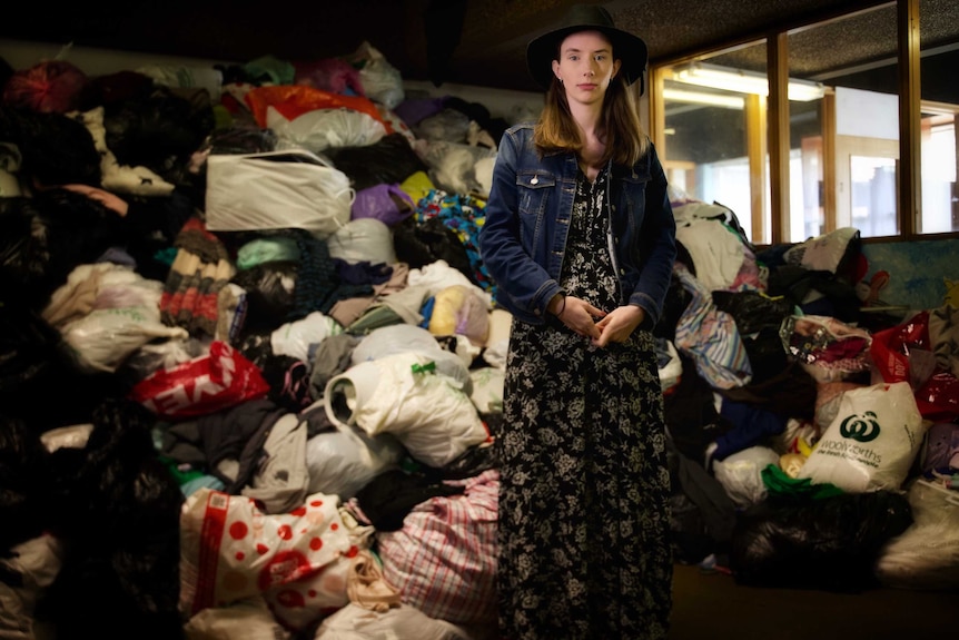Young woman in a dress, hat and denim jacket stands in front of a large pile of op shop donations.