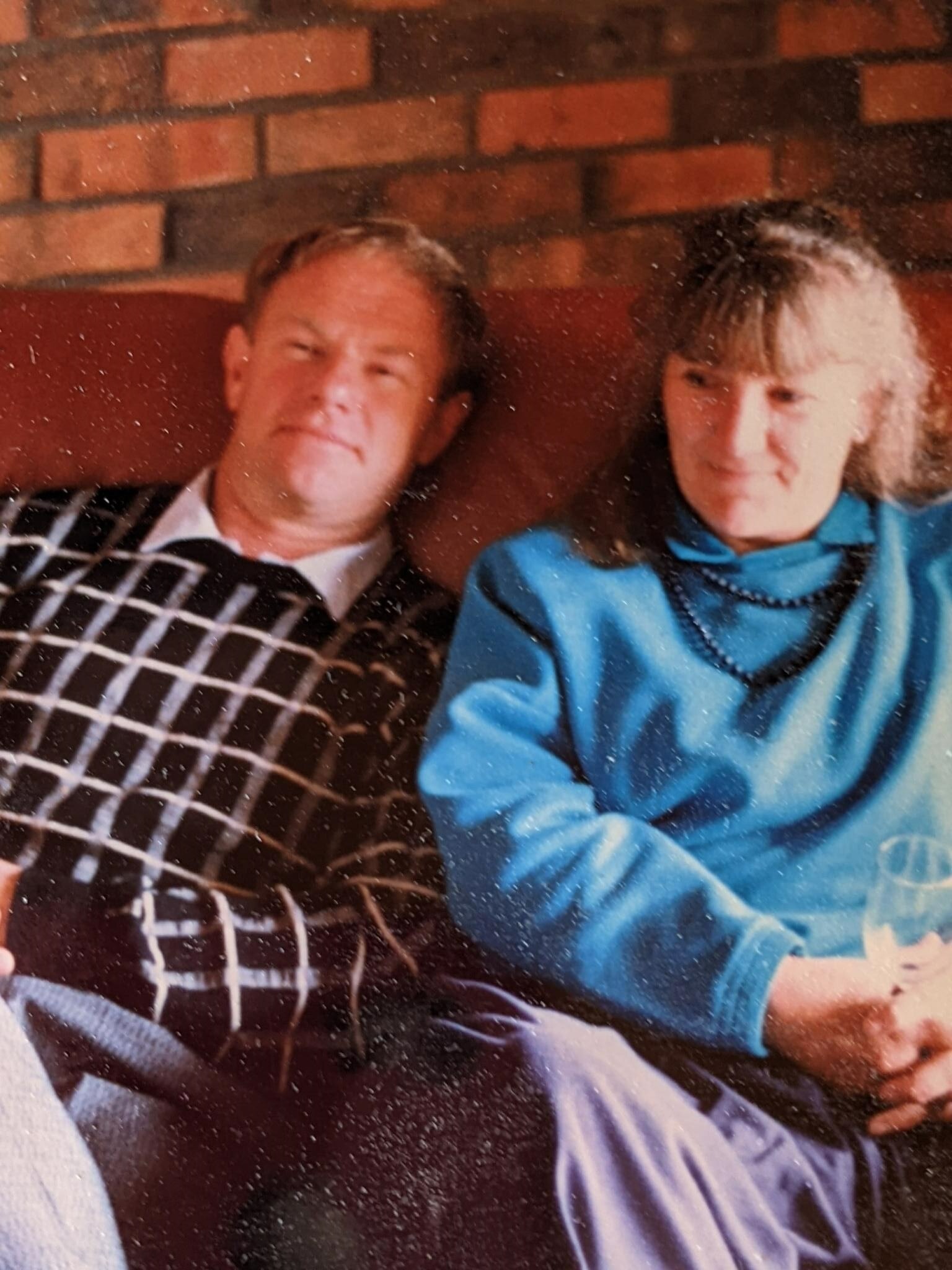   A photo from the late 80s of a couple sitting on a brown leather couch.  The woman is wearing a blue jacket and beaded necklace.