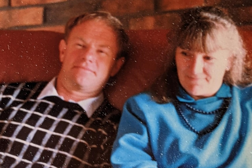  A photo from the late 80s of a couple sitting on a brown leather couch. The woman is wearing a blue jacket and beaded necklace.