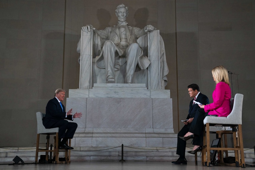 Donald Trump sits while speaking to reporters in front of a large statue of Abraham Lincoln