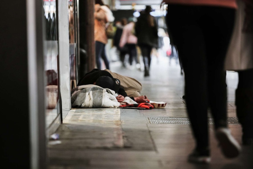 Homeless man sleeping on footpath with blankets as pedestrians walk past him in Elizabeth Street in Melbourne's CBD.