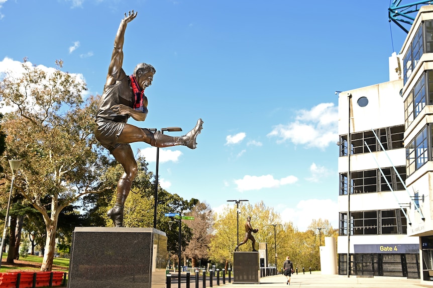 A large statue of AFL Legend Ron Barassi kicking a football stands outside at the MCG with a scarf hanging from it.
