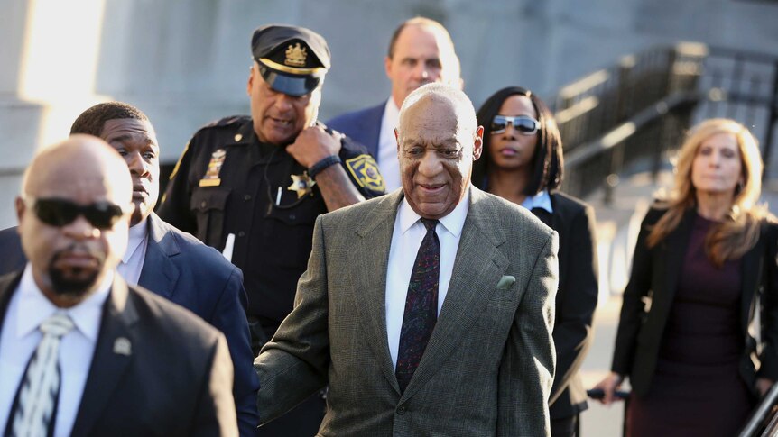 Bill Cosby walking wearing a suit and tie, surrounded by people in suits, as well as an official