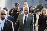 Bill Cosby walking wearing a suit and tie, surrounded by people in suits, as well as an official