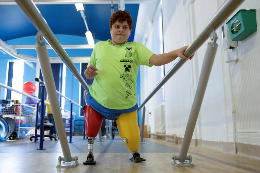 A young Turkish boy in a green T-shirt holds on to a pair of low bars as he walks on two brightly coloured prosthetic legs.