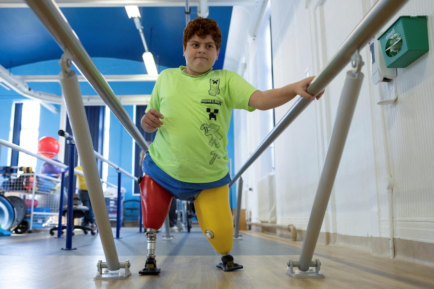A young Turkish boy in a green T-shirt holds on to a pair of low bars as he walks on two brightly coloured prosthetic legs.