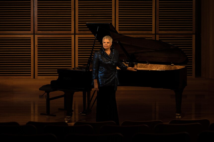 Deborah Cheetham Fraillon stands in front of a grand piano on the stage at the Sydney Conservatorium of Music's Verbrugghen Hall