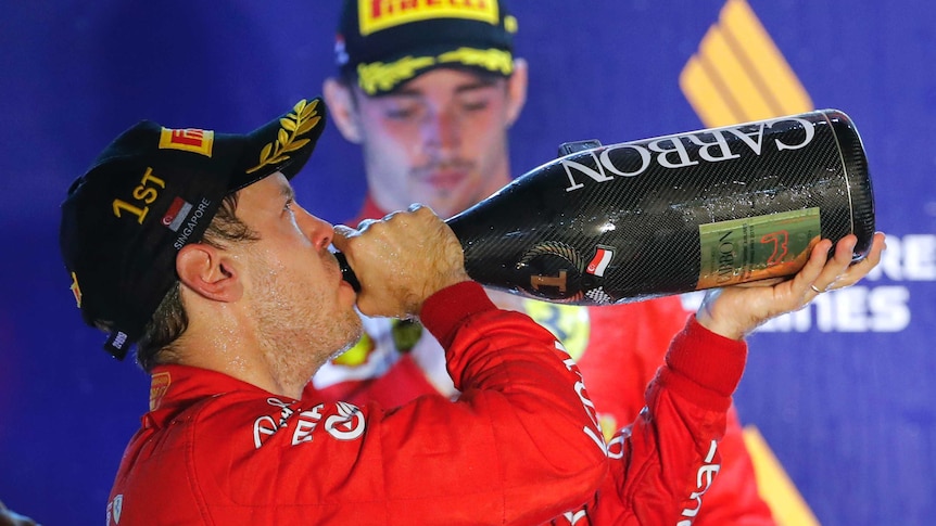 Sebastian Vettel leans back and drinks from an over-sized champagne magnum as Charles Leclerc looks downcast in the background