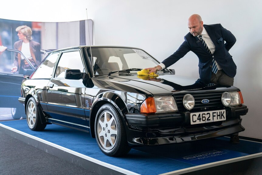 A man in a suit polishes a black Ford Escort RS Tourbo as it rests on a small carpet stage. 