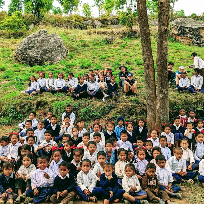 Lots of school students, most Nepalese, sit in a green field.