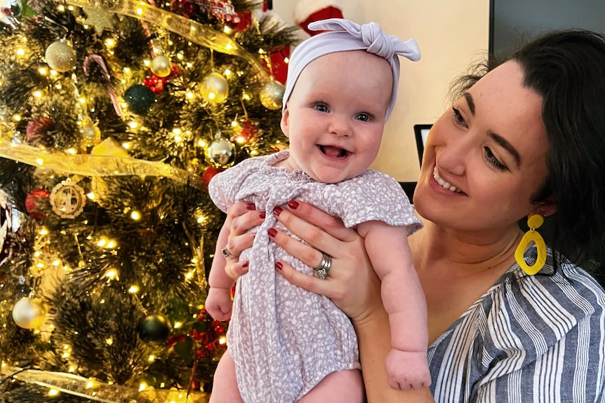 A woman holding a baby in front of a Christmas tree.