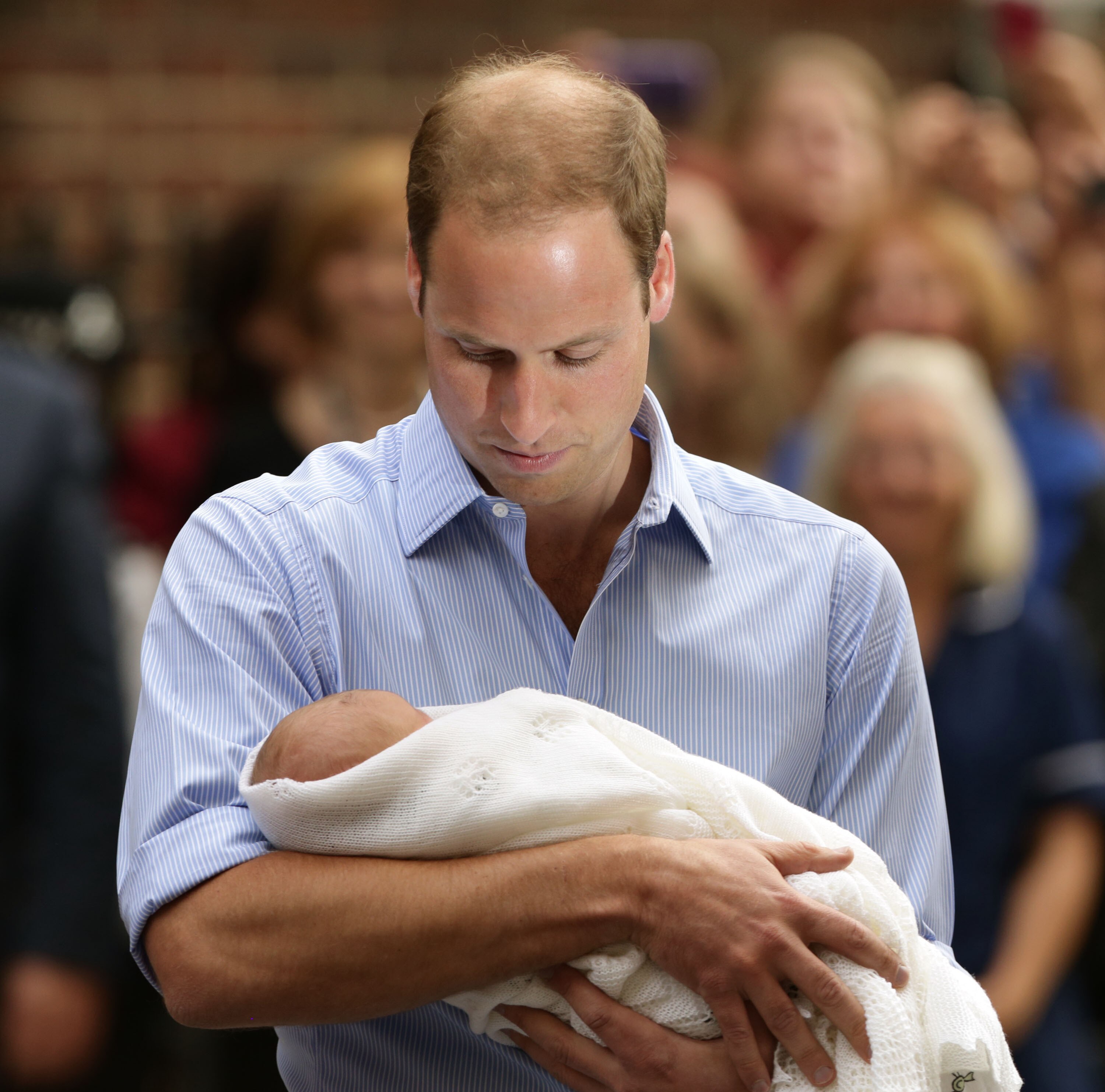 Prince William cradles the newborn baby George in his arms, looking tender, surrounded by a crowd.