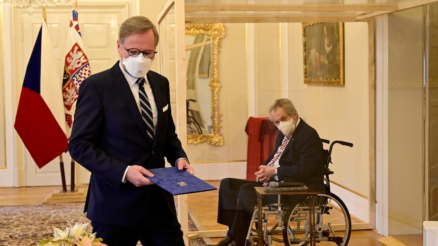 Man sitting in a wheelchair with a face mask in a glass box behind a man outside the glass box wearing a suit and mask 