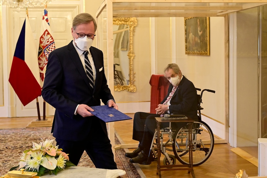 Man sitting in a wheelchair with a face mask in a glass box behind a man outside the glass box wearing a suit and mask 