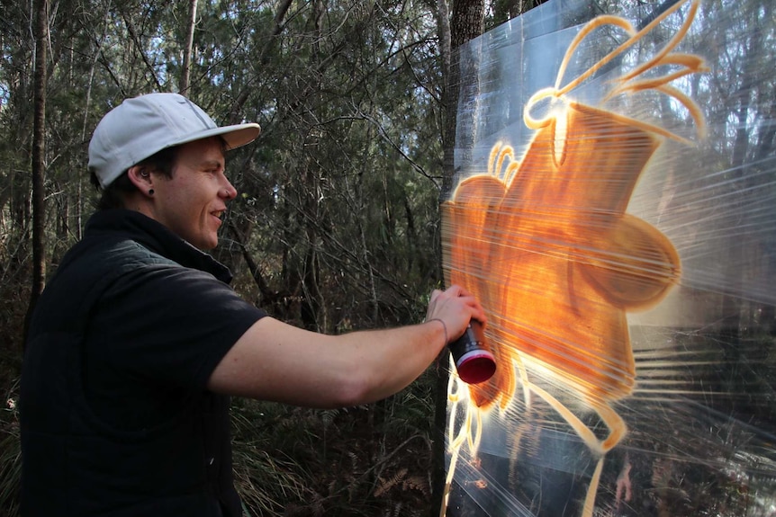 Tasmanian artist James Cowan spray painting a mural on plastic food wrap stretched between trees.