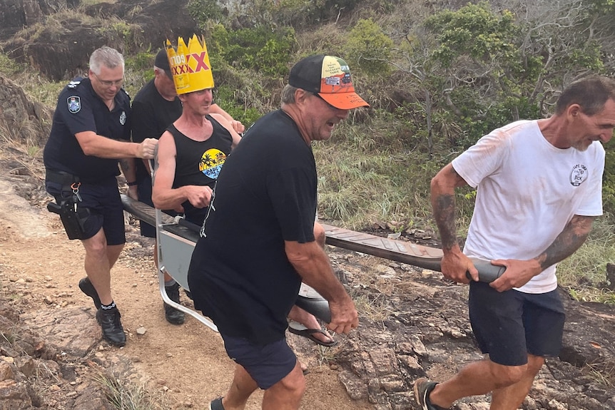 A man wearing a yellow cardboard crown is carried in a wooden chariot by four men over a rocky path.