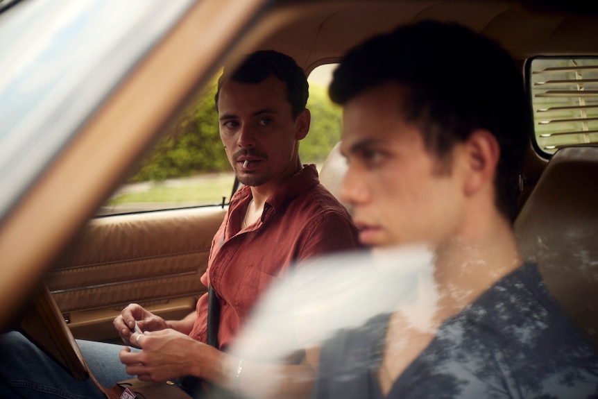 Two young men sit in the driver and passenger seat of a car. One is rolling a cigarette and looking intently at the other.