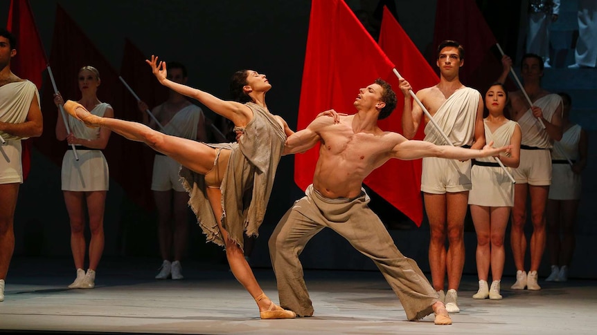 A male and female ballet dancer embrace on stage. Other dancers holding red flags stand behind them.