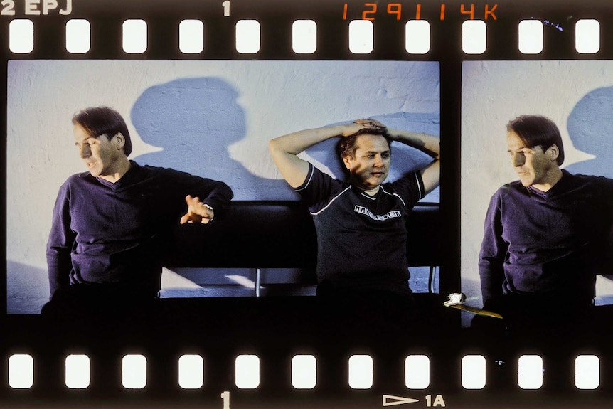 Two men sit on a leather bench chair against a white wall.