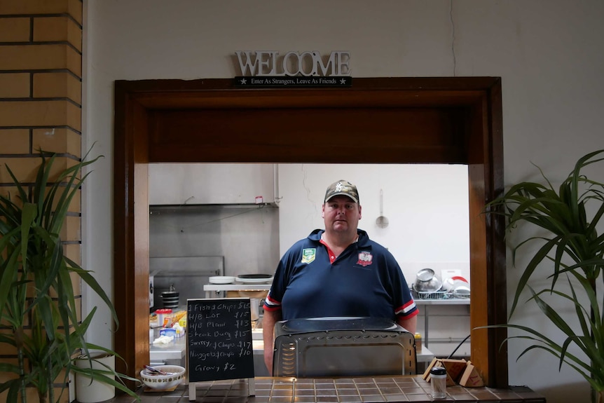 A man stands behind the dining room counter of an RSL club, with a kitchen behind him, and a Welcome sign on the wall above.